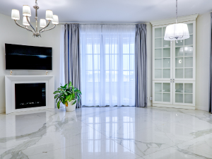 Bright living room with marble floor, wall TV above a white fireplace, a potted plant, and glass-front cabinets near large windows with sheer curtains.